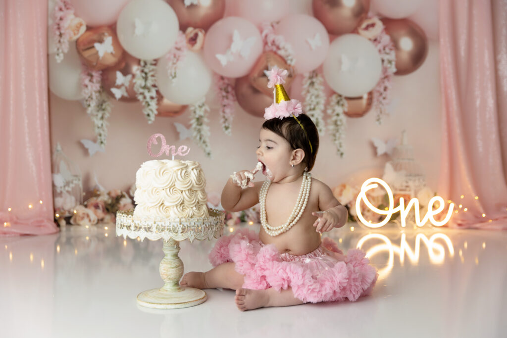 1st Birthday girl wearing a pink tutu & gold party hat about to put cake icing in her mouth during her 1s birthday photo shoot.