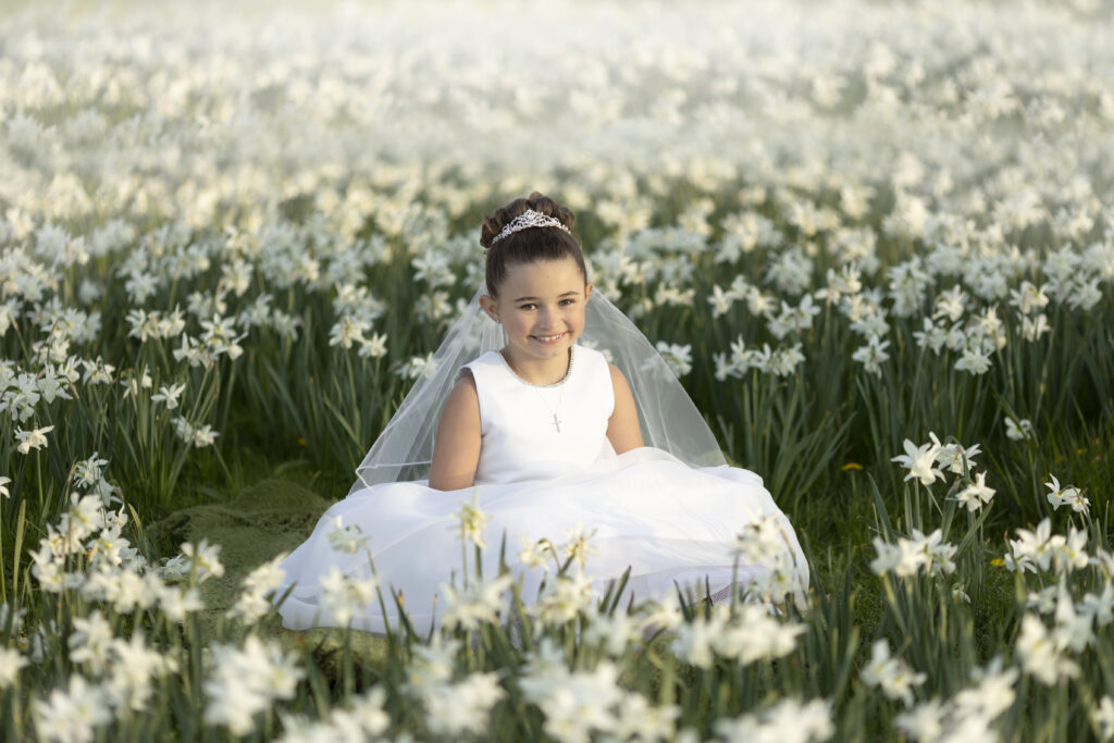 Young girl in First Holy Communion Dress sitting in a field of white flowers in an NJ park.