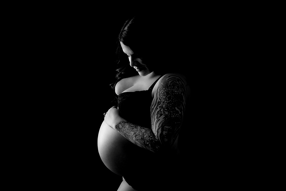 Black & white image of an expecting mother to be wearing a black bra looking down at her growing belly.