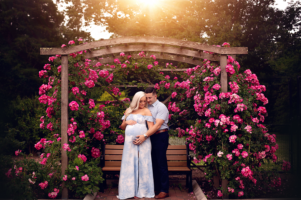 husband & wife expecting their first baby, take maternity photos under a rose covered arch in an NJ park.