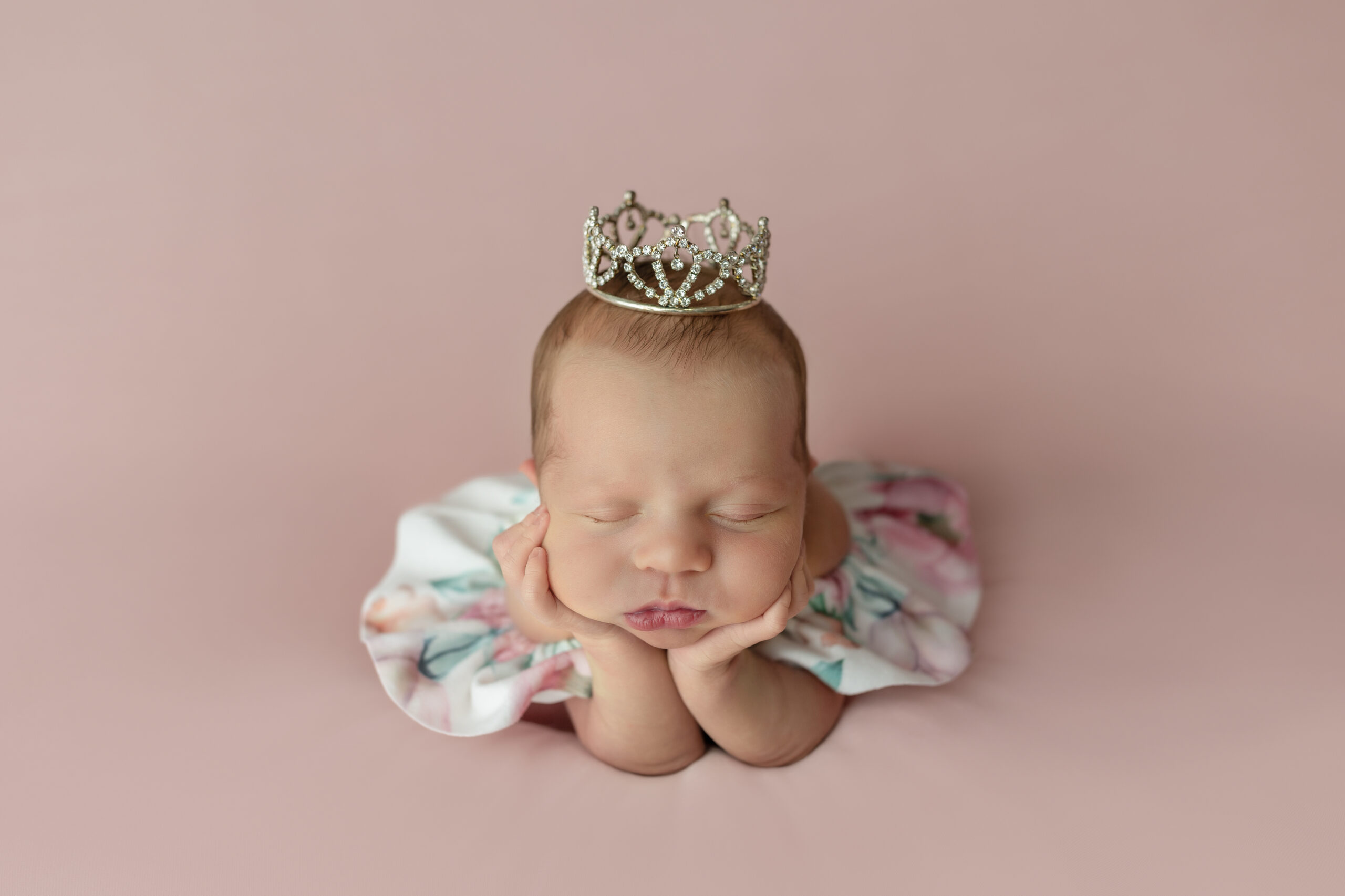 Newborn girl wearing a floral romper & princess crown in froggy pose.
