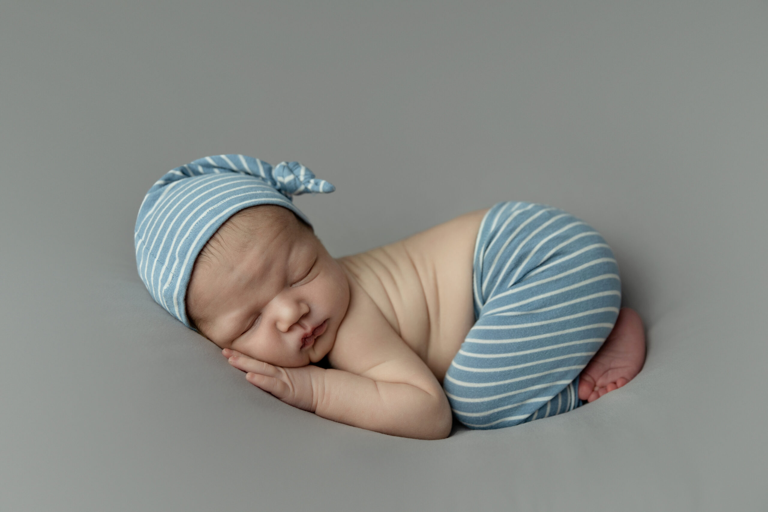 Newborn baby boy wearing blue striped pants & matching hat sleeping on his belly all squished up.