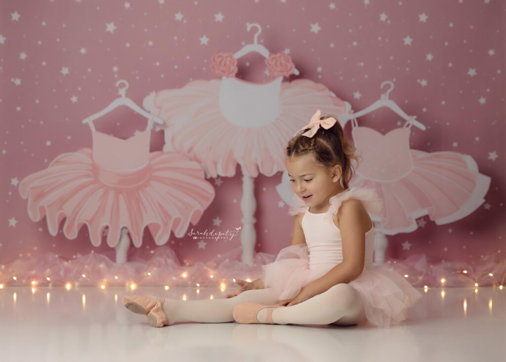 Little girl in pink leotard stretching for photos on a pink tutu background.