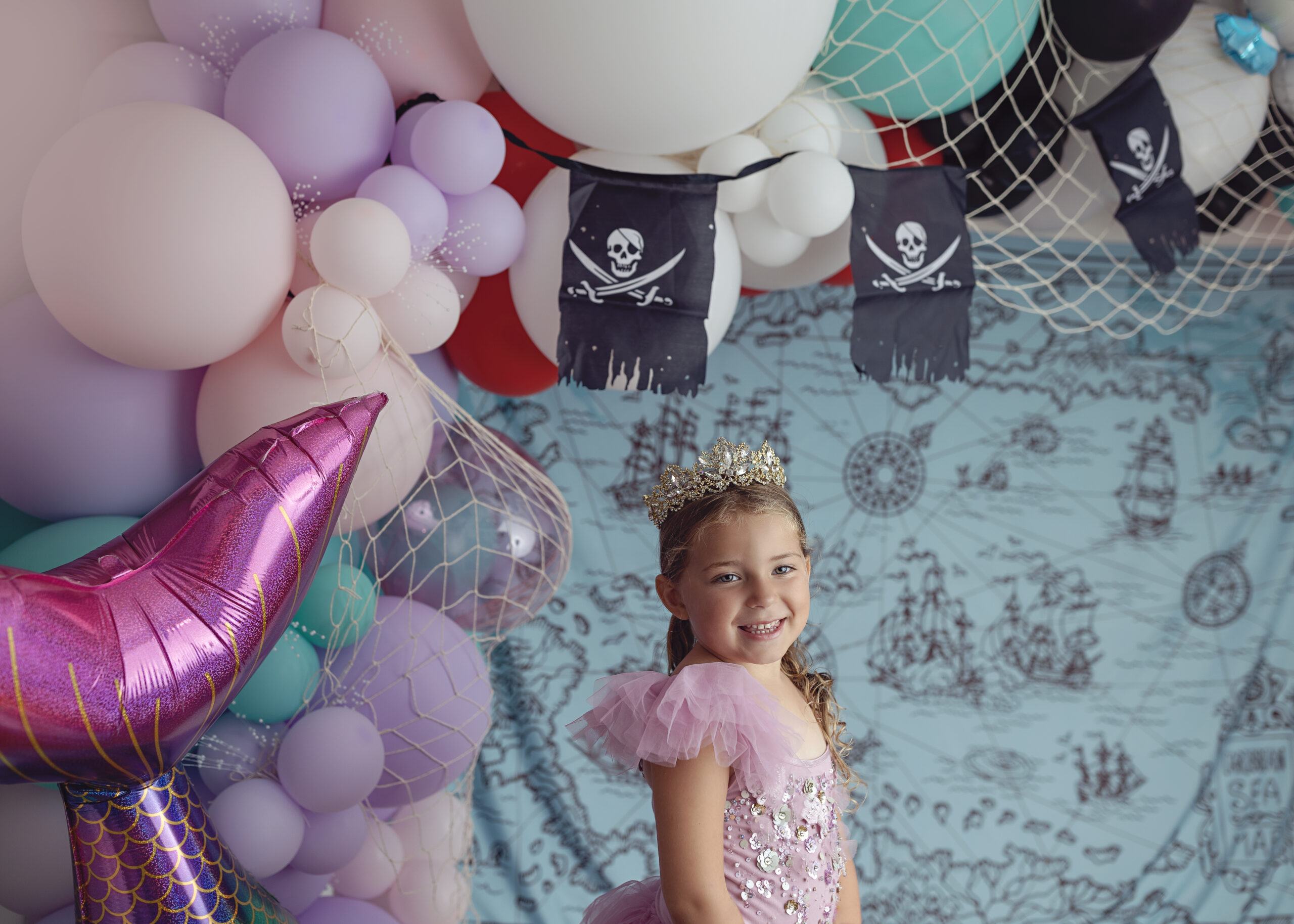5th birthday girl wearing fancy dress & crown in front of pirate & mermaid themed balloon arch and map backgrounds