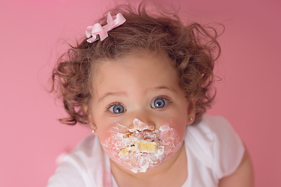 first birthday girl eating a smash cake with cake all over her face on a pink background