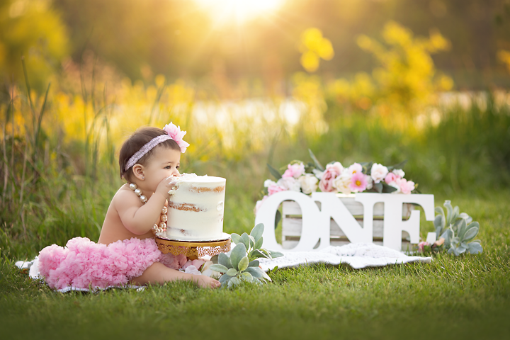 1st birthday girl in a pink tutu &. headband with pearls around her neck trying to eat her smash cake outside with a ONE sign & pink flowers in background