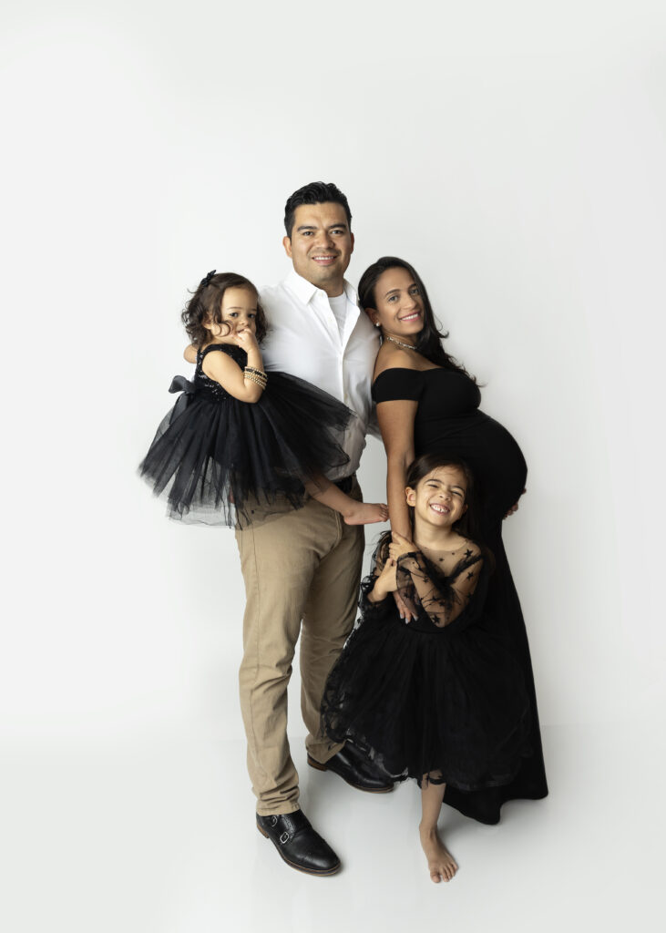 Pregnant mother, father, & two small daughters dressed in black taking photos on a white background.