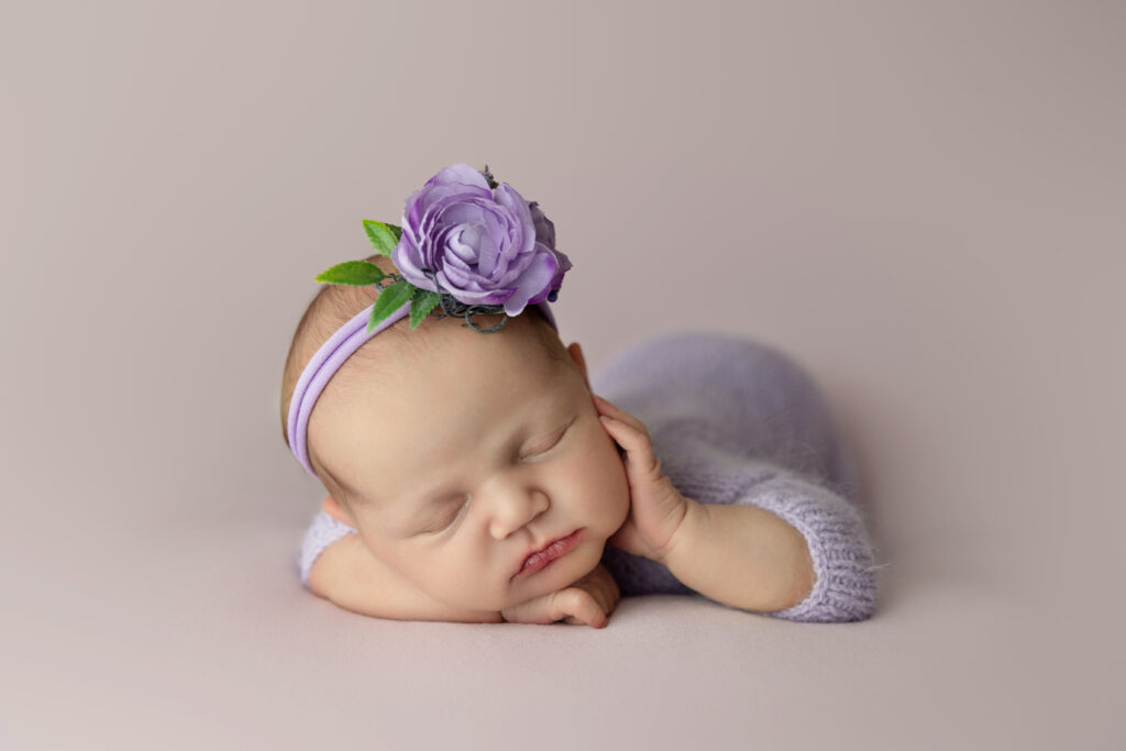 newborn baby girl in lavender romper with purple flower in her air sleeping with hand on cheek