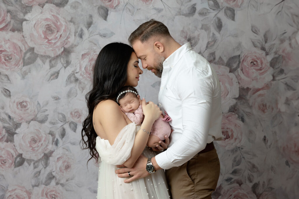 new mom & new dad holding baby girl in pink lace one a pink & grey floral background
