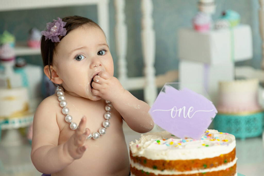 baby girl with pearl necklace, purple flower in her hair wating cake in font of a backer background and ONE on her smash cake