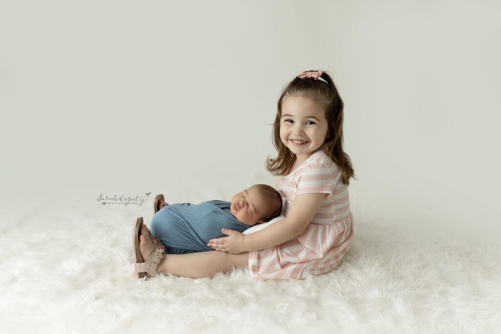big sister wearing white & white striped shirt holding newborn baby brother swaddled in blue on a back fur rug