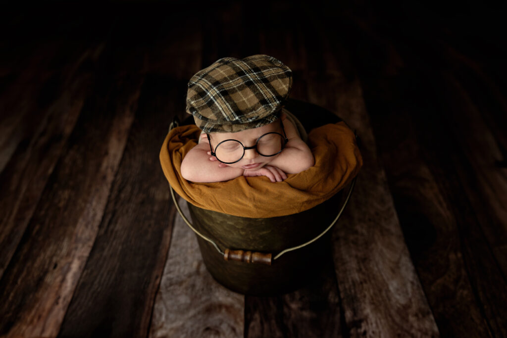 newborn baby boy with old fashion hat & glasses sleeping in a bucket on a dark brown wood floor