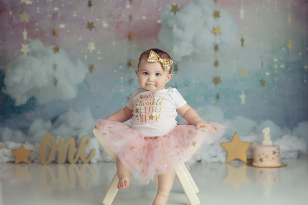 baby girl in pink tutu with gold stars on a pastel sky backgrounds with one and a cake in background